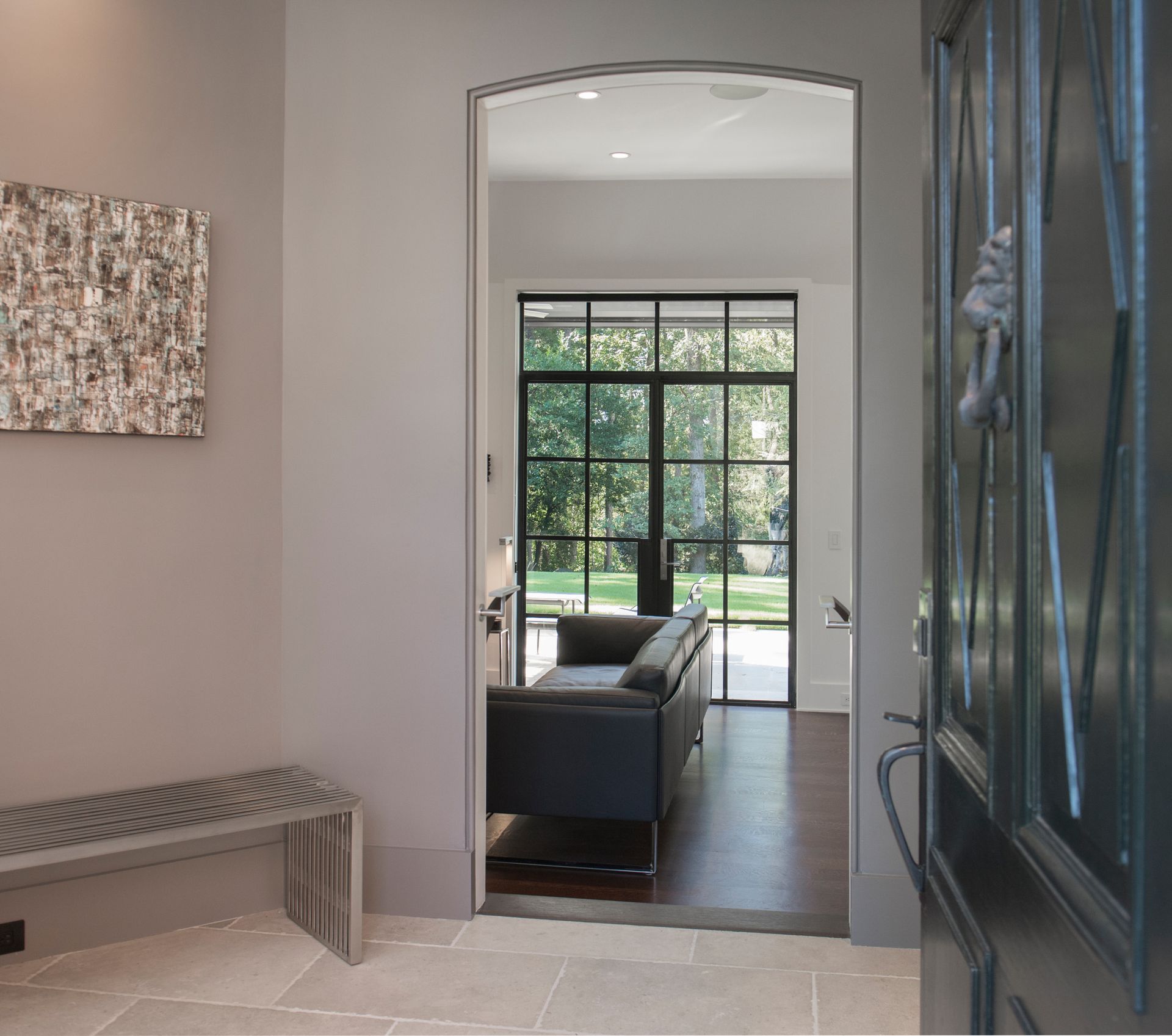 Entryway: gray walls, bench, artwork. View through an archway of living room with black sofa and door to backyard.