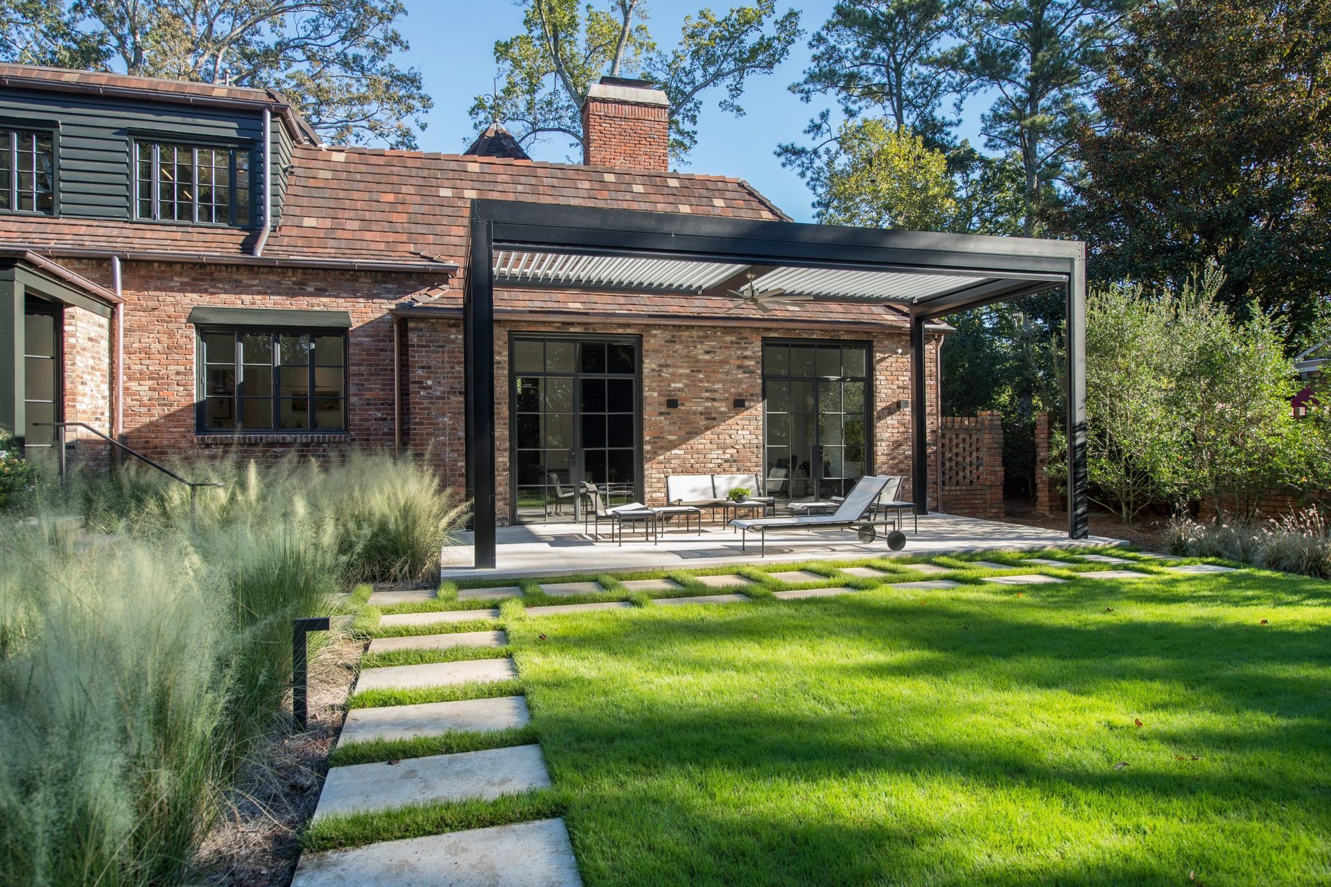 Backyard patio with pergola, brick house, green lawn, stone walkway, tall ornamental grass.