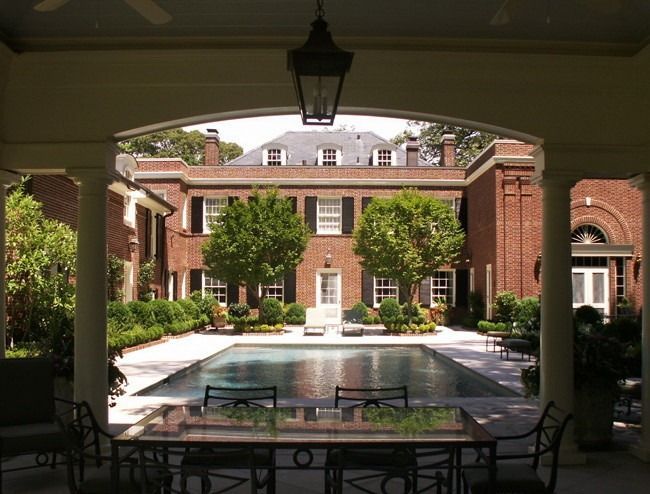 Pool and patio of a large brick home, viewed from under a veranda with columns and a hanging lamp.