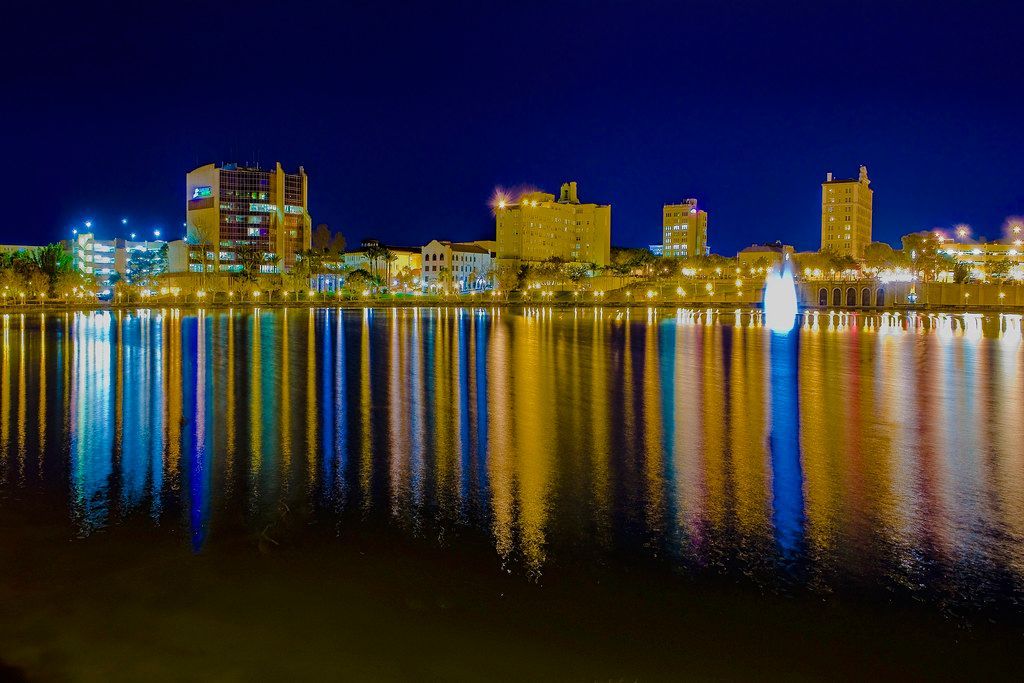 Night view of a city skyline with buildings reflecting colorful lights on the water.