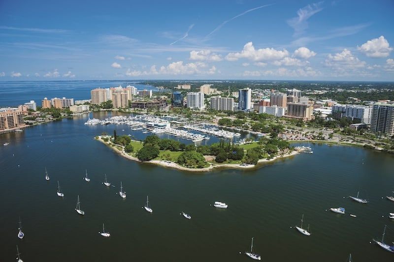 Aerial view of Sarasota Bay with sailboats, marina, and downtown skyline on a sunny day.