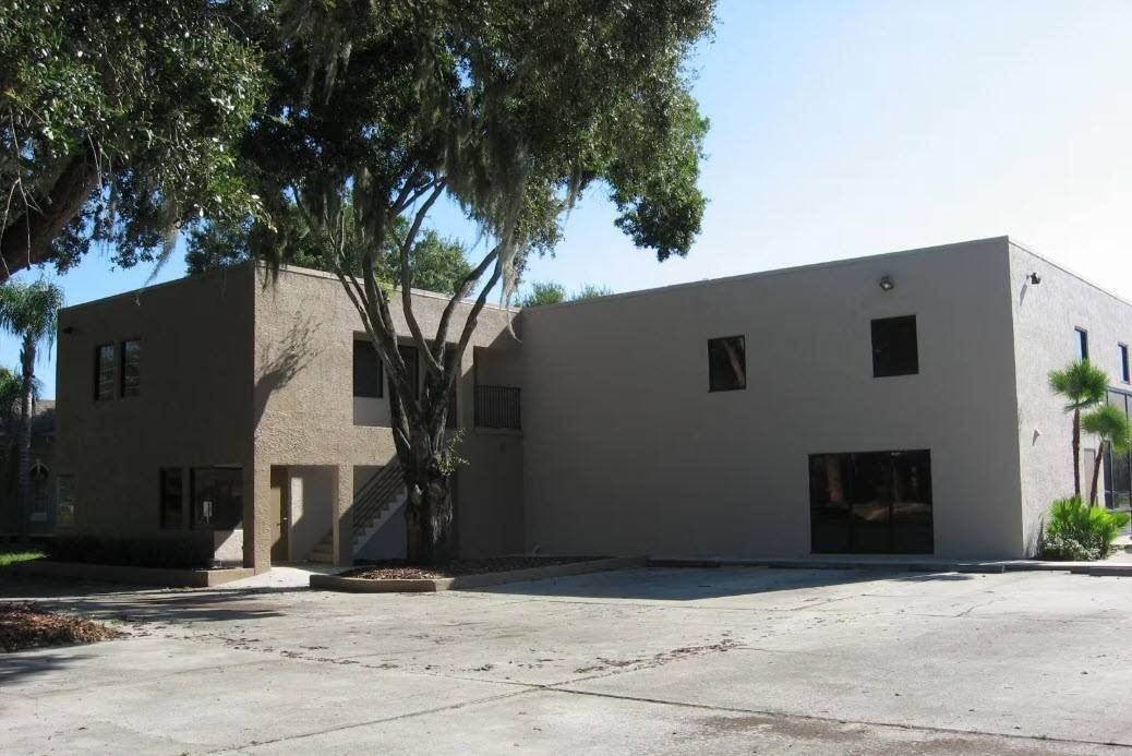 Modern two-story beige building with trees casting shade and a paved parking area in front