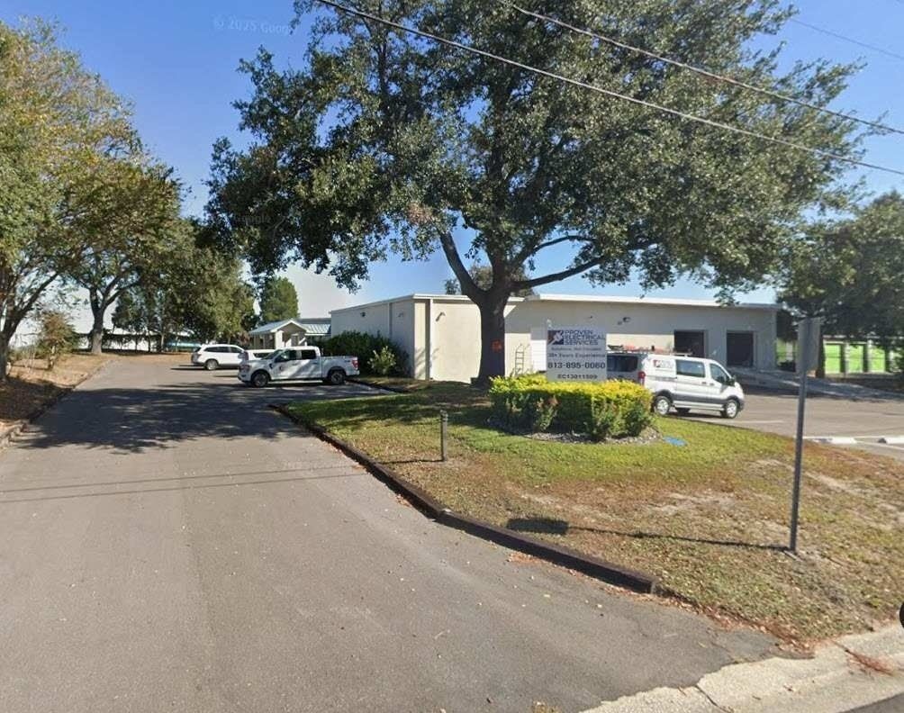 Quiet street beside a small industrial building with parked cars, trees, and a sign near the entrance.