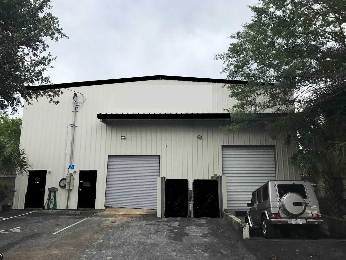 White industrial warehouse with two roll-up doors, parked vehicles, and trees outside