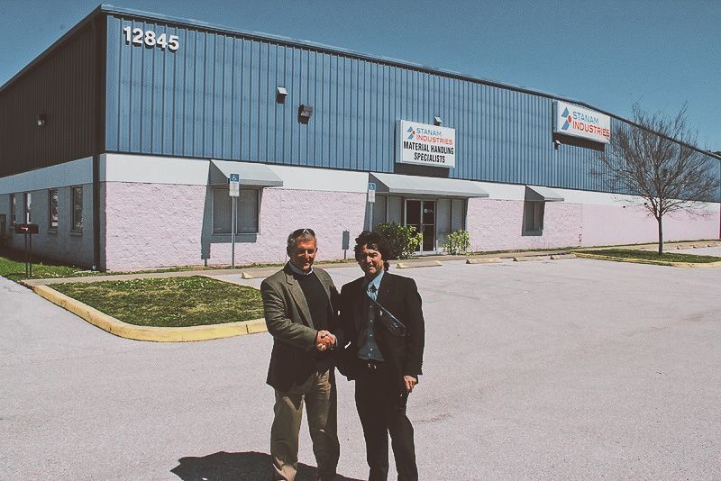 Two people stand outside a commercial building with signs on the front and a large parking lot.