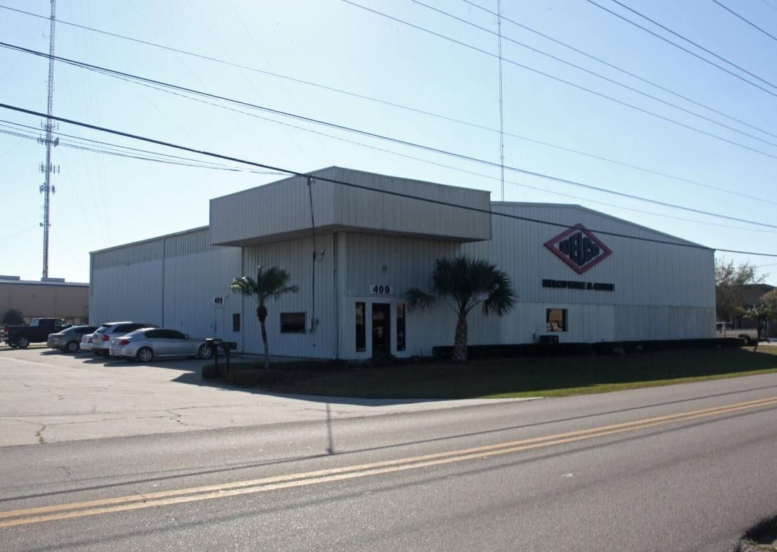 Large white industrial building with parked cars, palm trees, and roadside signage under a clear sky