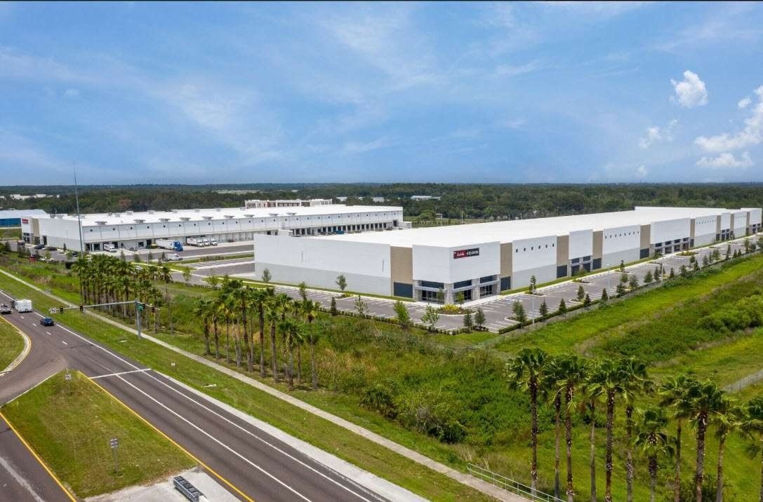 Aerial view of a large white industrial complex beside a road with palm trees and green fields under a blue sky