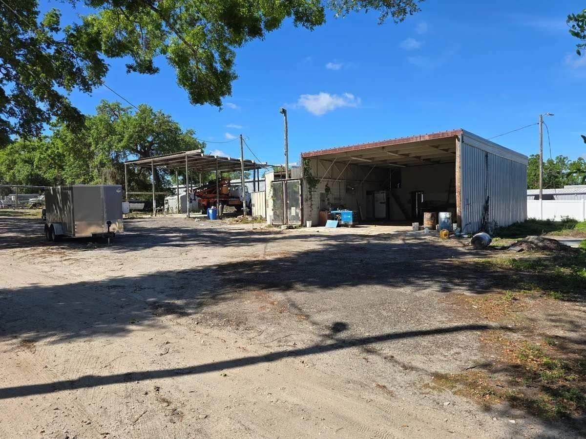 Rural yard with a large metal shed, a trailer, and sparse trees under a bright blue sky.