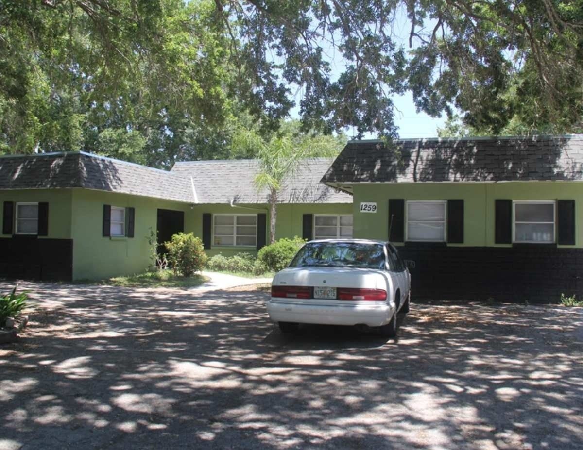 Single-story house with green siding, black shutters, and a parked silver car in the driveway under trees.