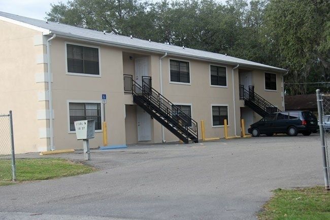 Two-story beige apartment building with exterior stairs, parked car, and chain-link fence in front