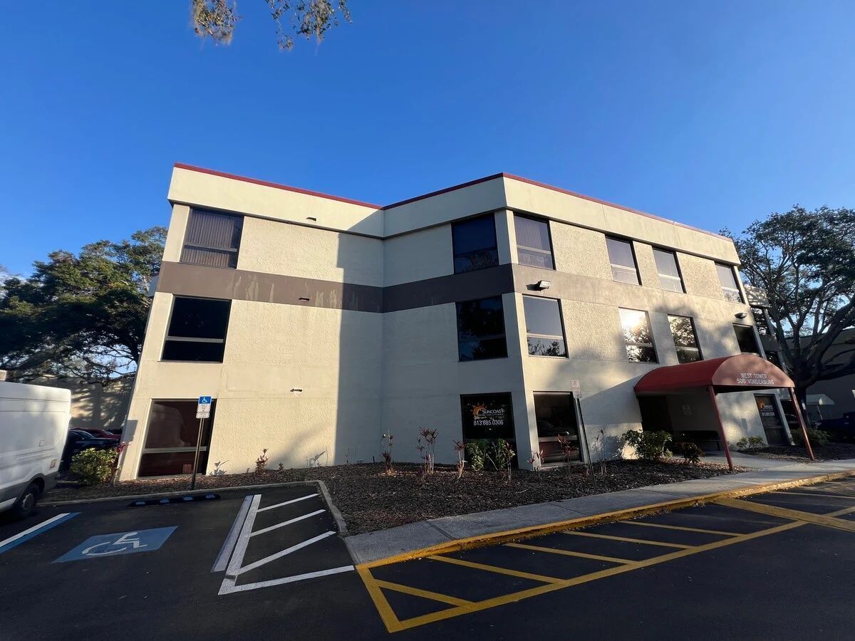 Modern two-story beige office building with dark windows and a red awning, viewed from a parking lot.