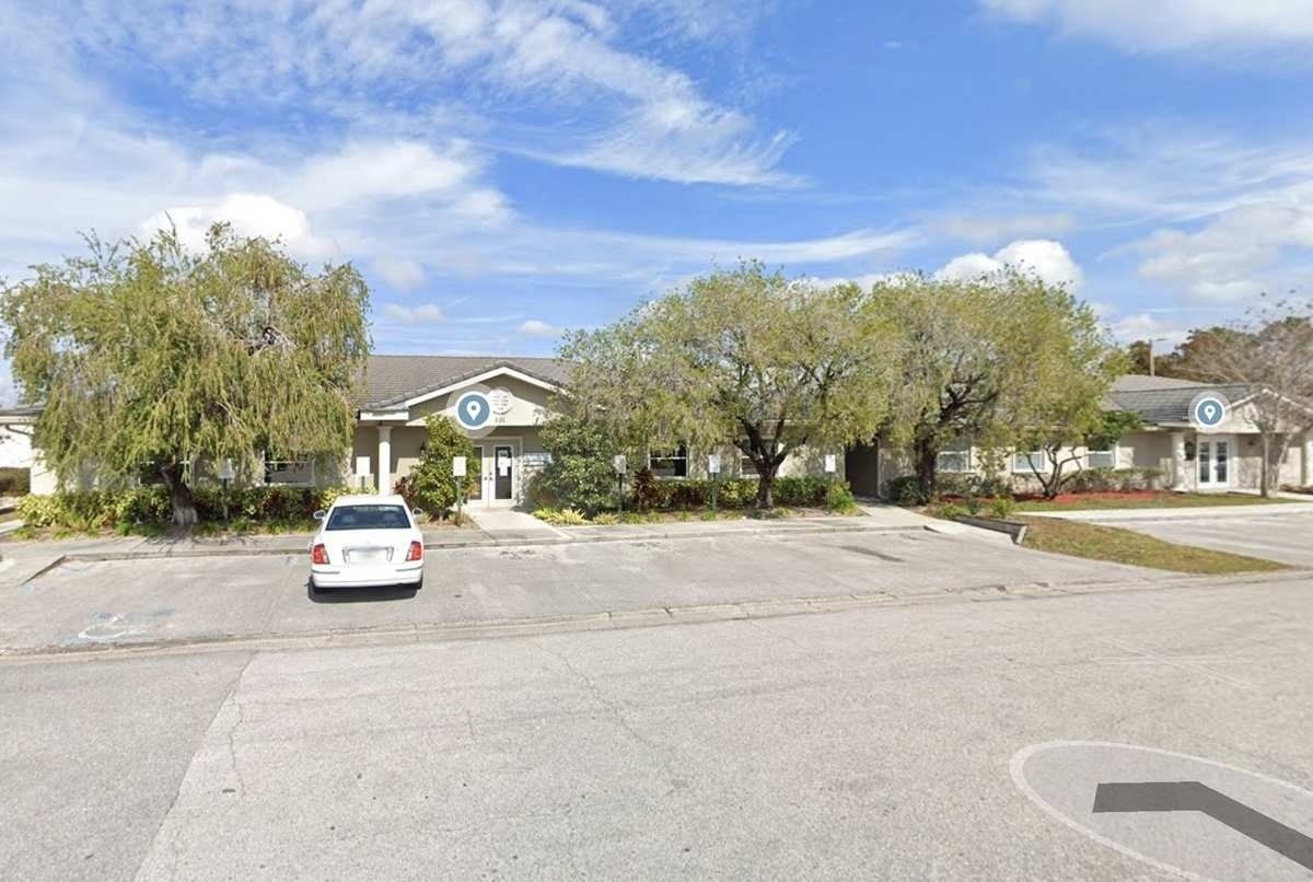 Parking lot in front of a low, beige building with trees and a white car parked near the entrance.
