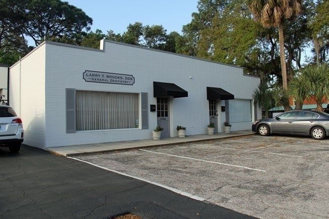 Small white commercial building with parked cars and a paved lot surrounded by trees