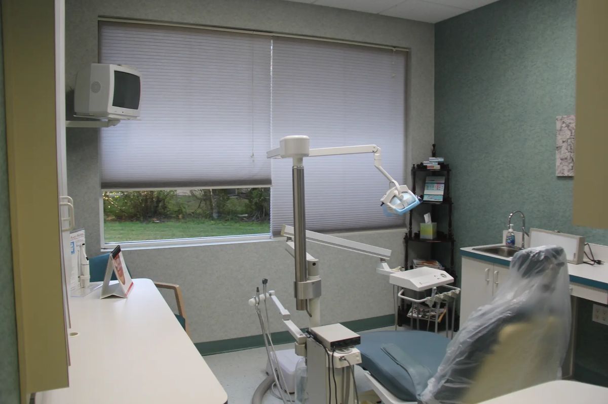 Empty dental exam room with chair, overhead light, window blinds, and medical equipment