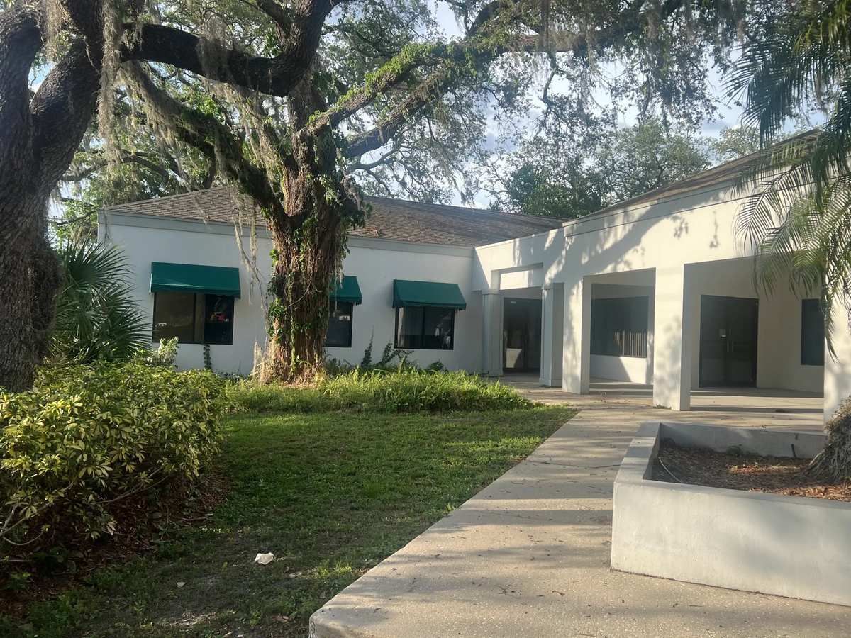 Courtyard with white buildings, green awnings, large trees, and a concrete walkway in sunlight
