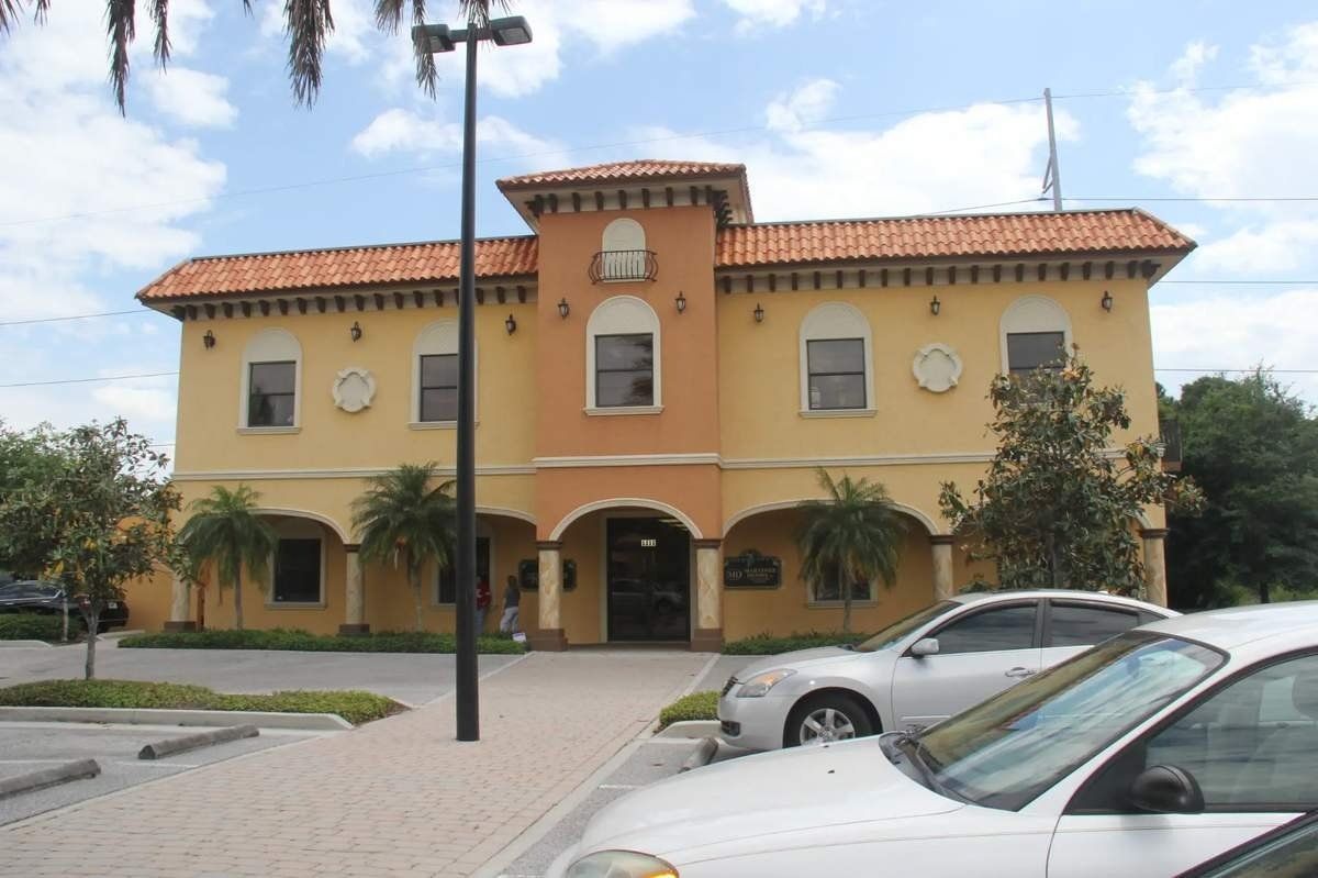 A two-story, yellow Spanish-style building with a terracotta tiled roof, arched entrance, and parking lot in front.