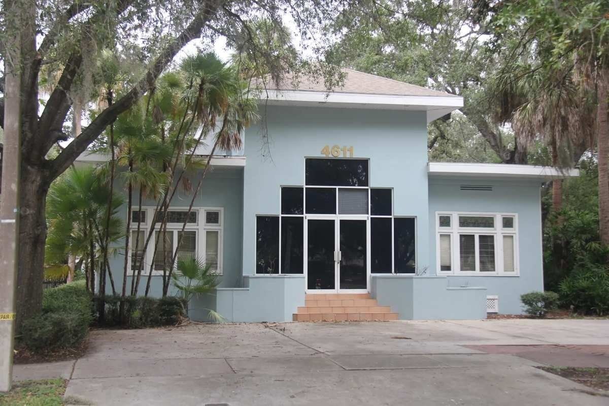 Small blue building with glass entrance, trees, and a paved driveway.