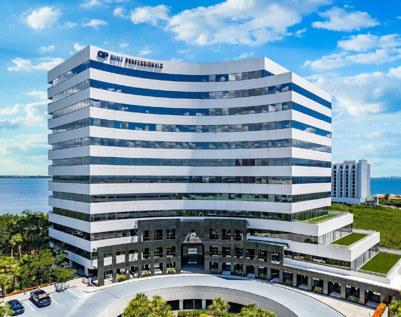 Modern glass office building with blue-tinted windows beside greenery and the ocean