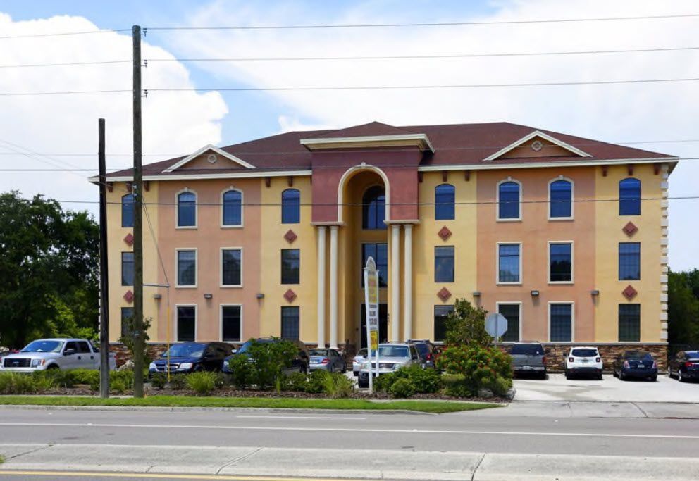 Three-story beige and red apartment building with blue windows beside a road under a cloudy sky