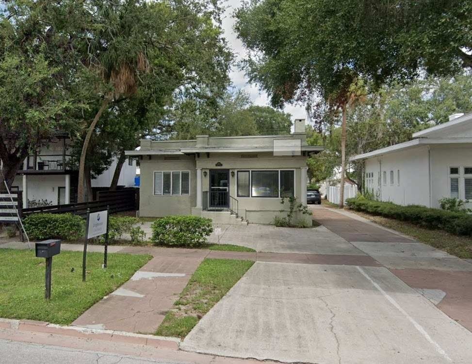 Small beige house with a front porch and driveway, surrounded by trees and a lawn on a quiet street.