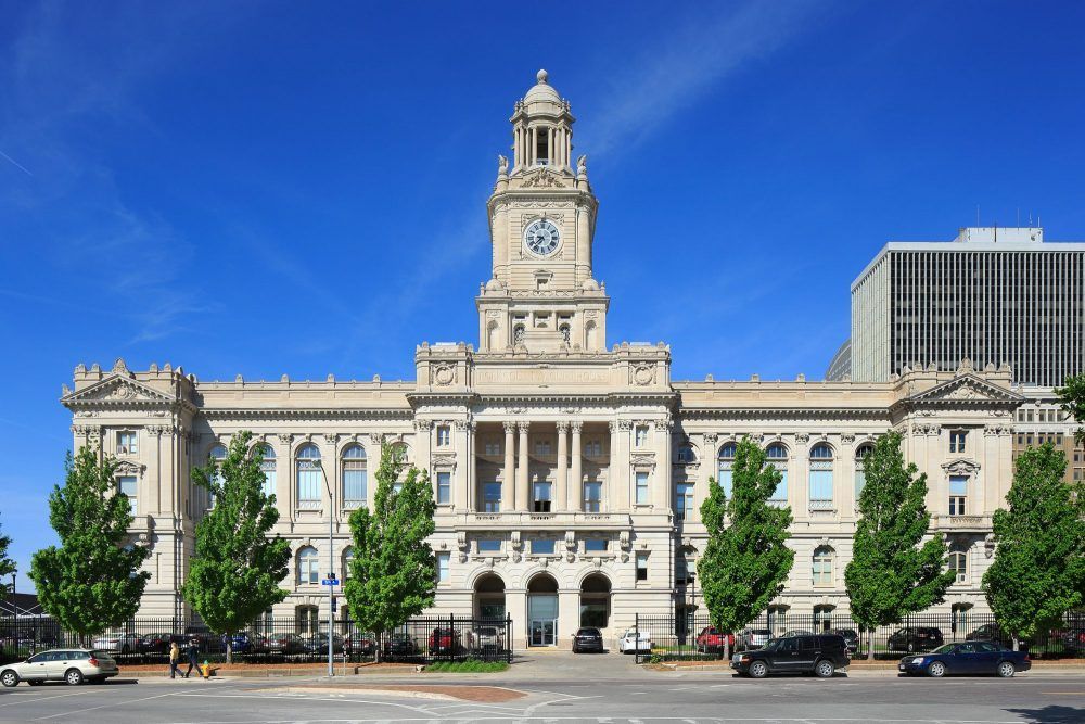 Beige building with clock tower and trees against a blue sky; cars and pedestrians in front.