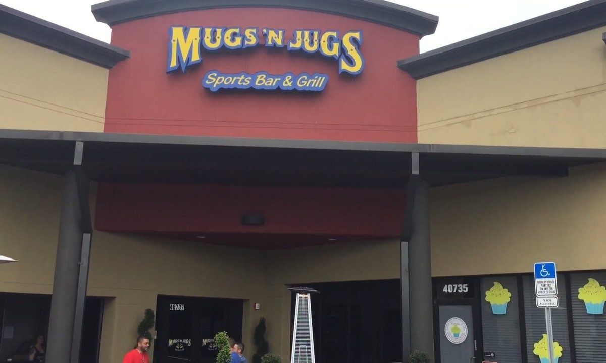 Storefront sign for “Mugs N Jugs Sports Bar & Grill” on a beige building with a covered entrance