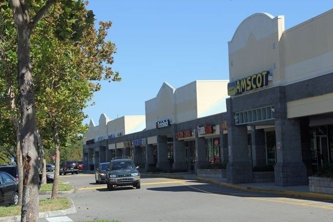 Small strip mall storefronts on a sunny street with parked cars and trees