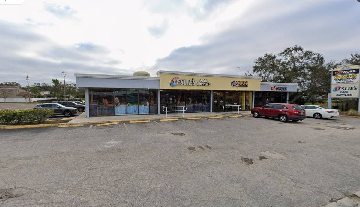 Small roadside strip mall with a yellow storefront sign, parked red car, and empty gravel lot.