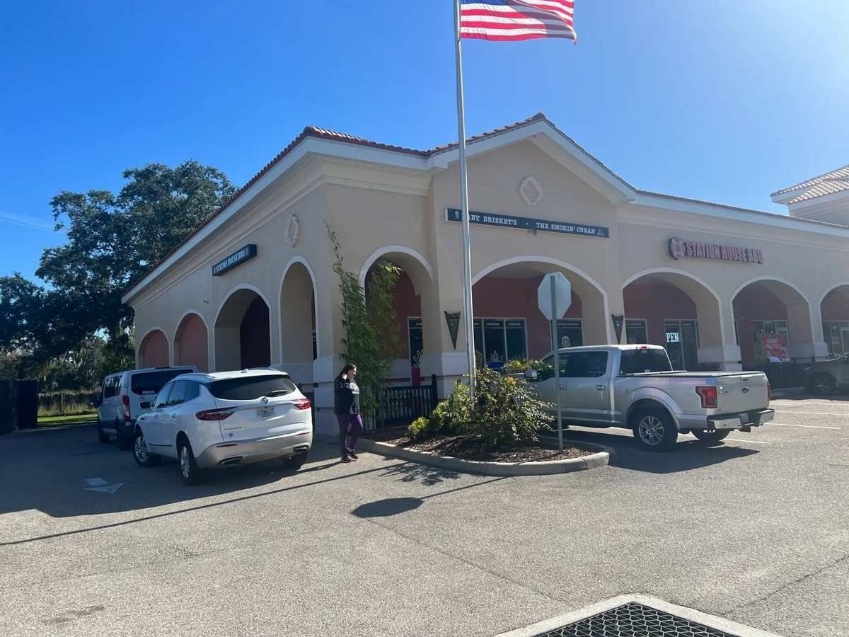 Small beige storefront with arched windows, parked cars, and an American flag waving under a blue sky