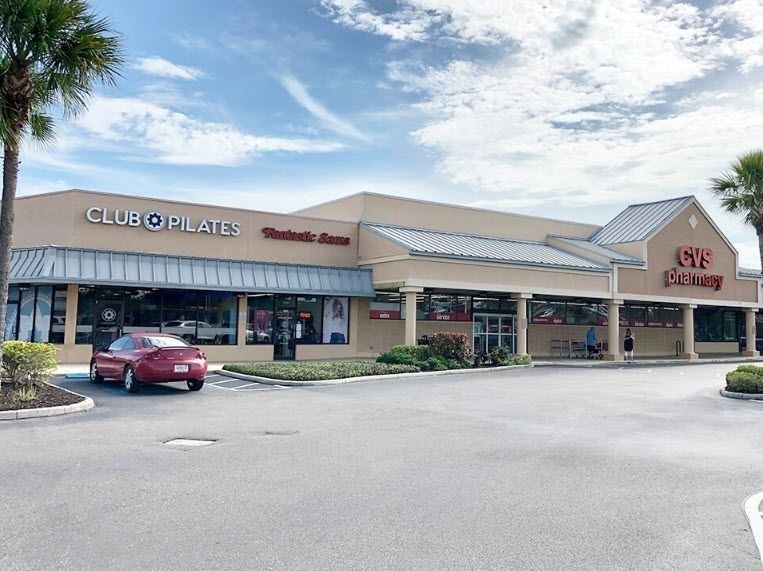 Strip mall storefronts under a cloudy sky, with a red car parked in front.