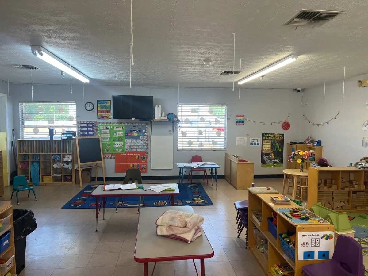 Empty preschool classroom with colorful rugs, tables, shelves, and toys under bright windows