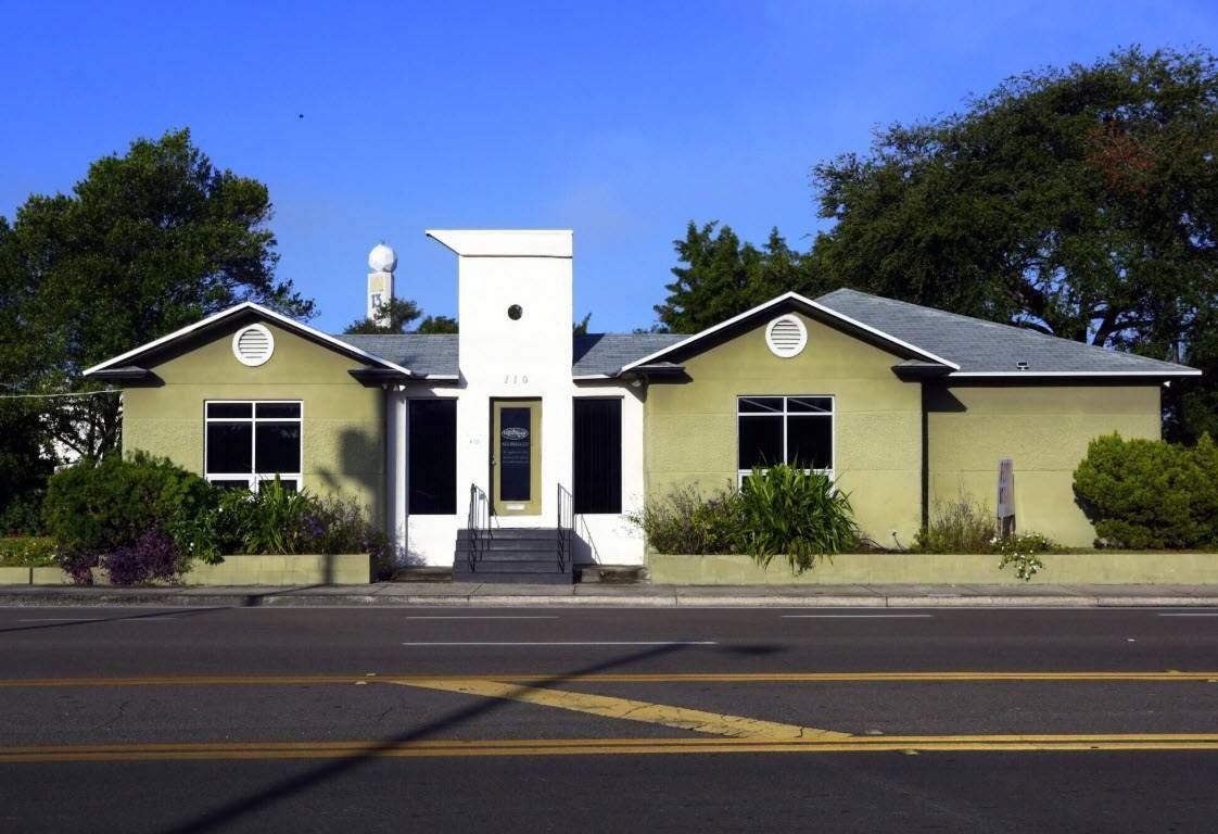 Small beige house with a white central entryway, black roof, and trees in front by a road