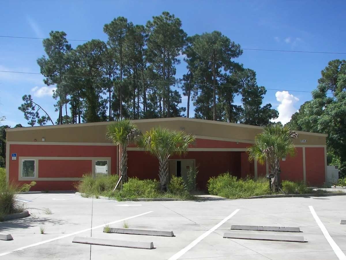 Single-story red building with white trim and a parking lot in front, under a blue sky with trees behind.