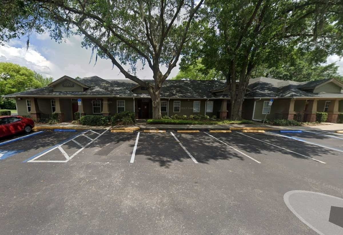 Single-story brick office building with shaded parking spaces and large trees in front.