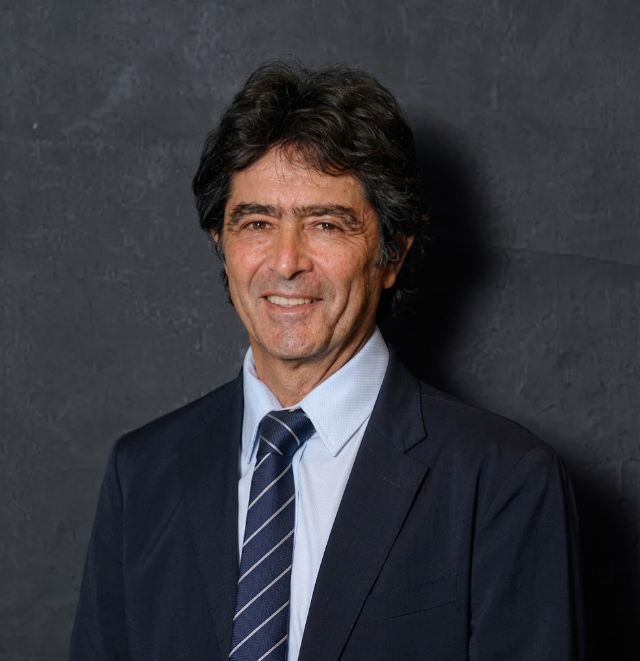 Man in suit smiles. Dark hair, blue tie, against dark background.