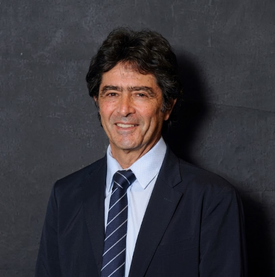 Man in suit smiles. Dark hair, blue tie, against dark background.
