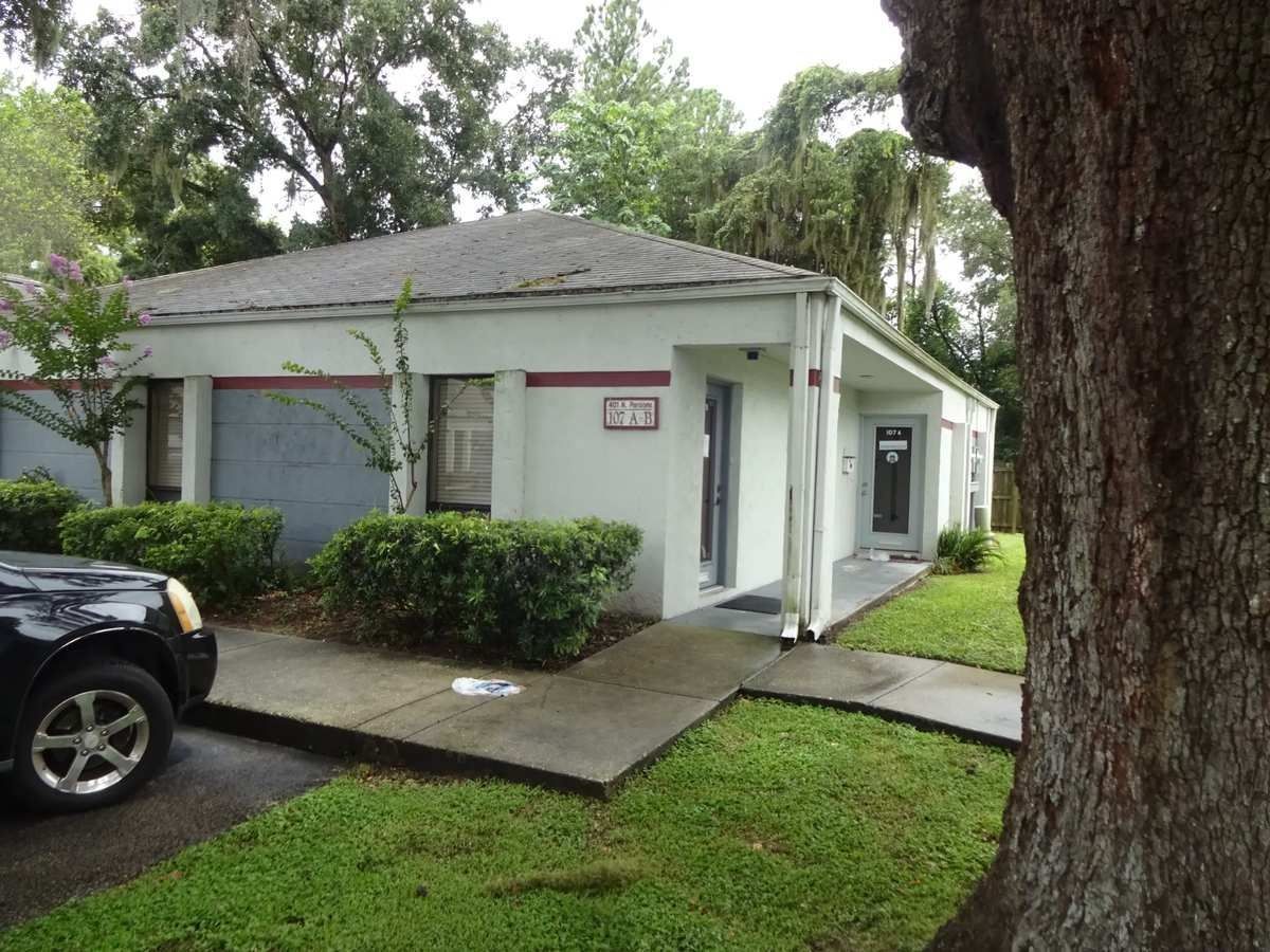 Small white house with a front porch and walkway, surrounded by trees and shrubs on a rainy day
