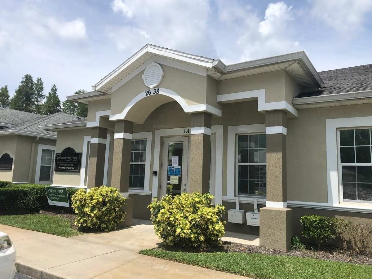 Single-story beige building entrance with white trim, arched doorway, windows, and shrubs along the sidewalk