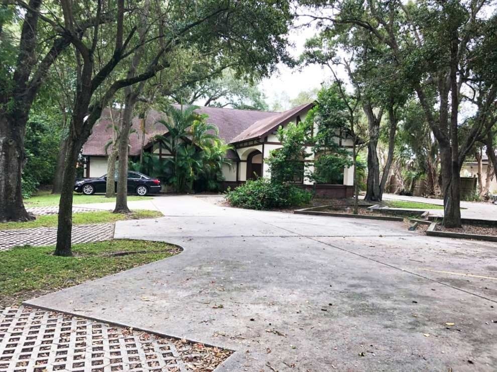 Tree-lined driveway leading to a house with a red roof and parked cars.