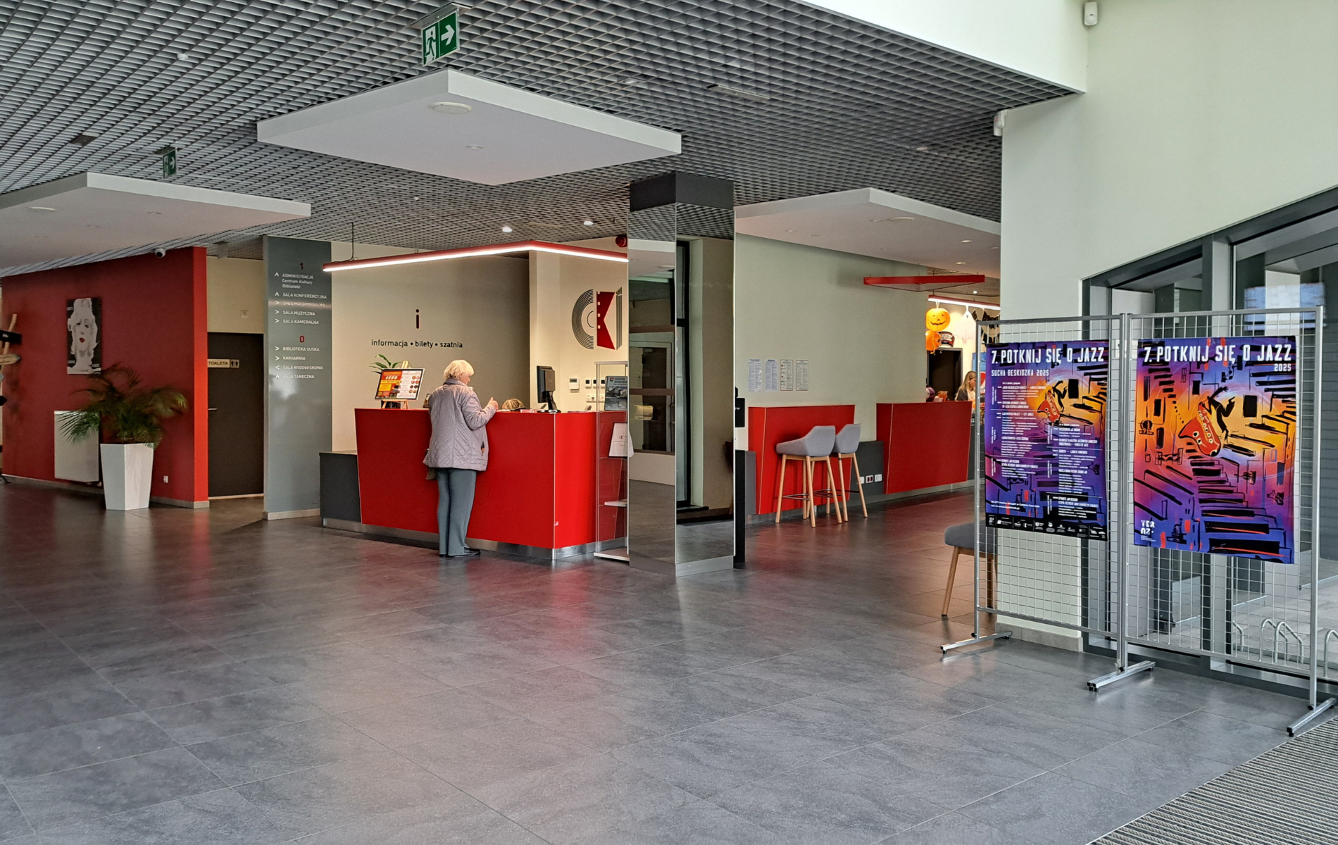 Interior lobby with red counters, a person at the reception desk, posters, and a gray floor.