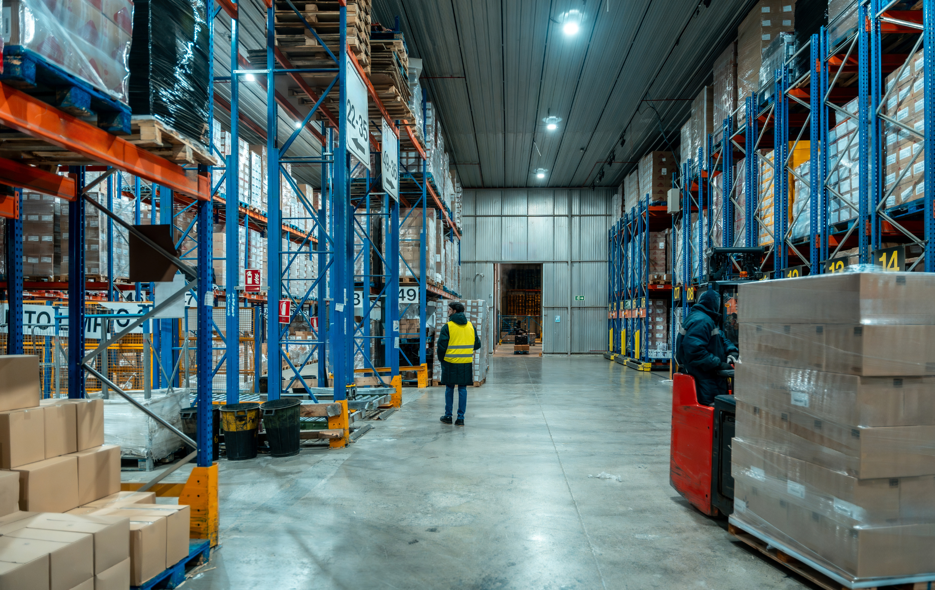 Warehouse interior with high shelving stacked with goods. Worker in safety vest walks toward a forklift.