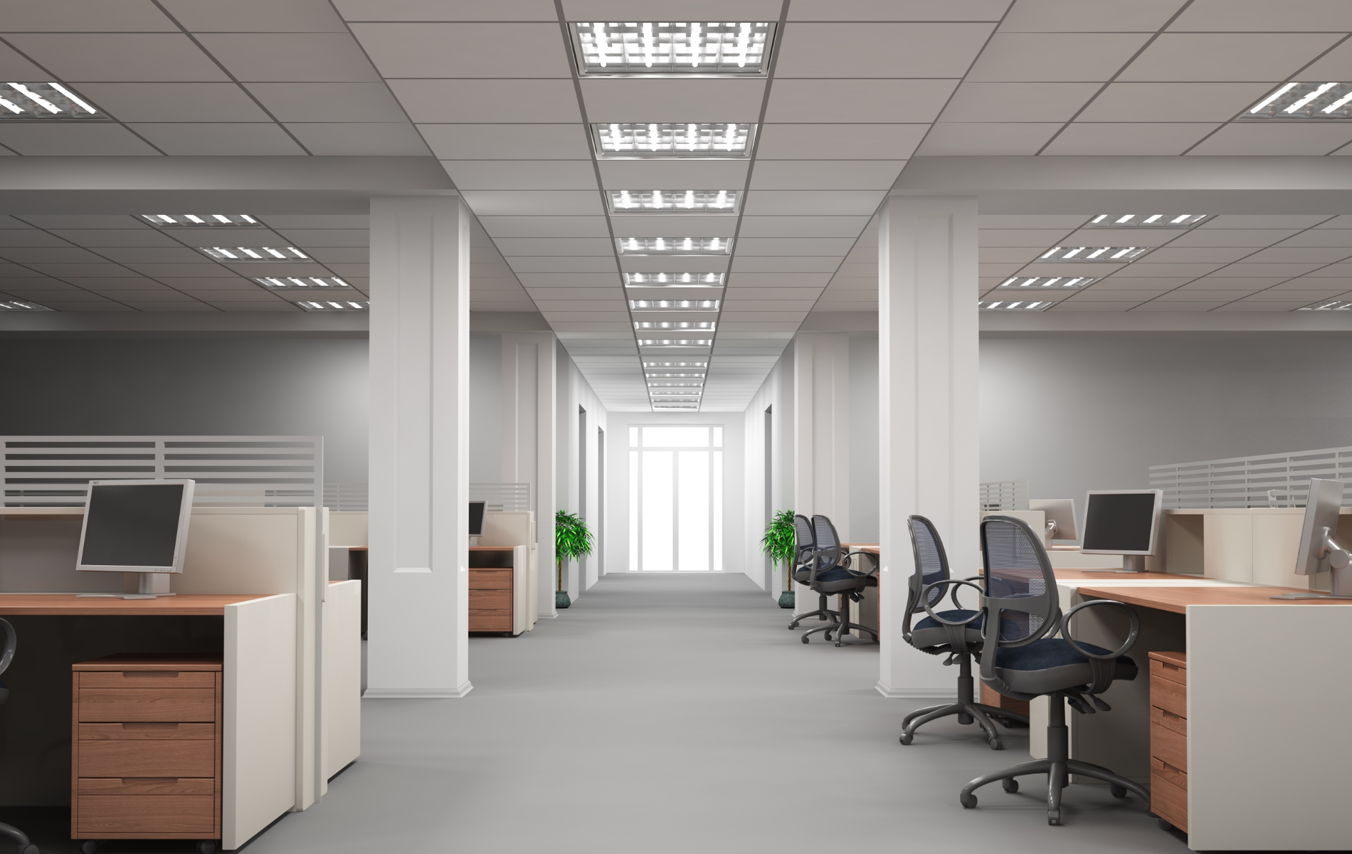 An empty office with rows of cubicles, desks, chairs, and overhead lighting. The hallway leads to a bright window.