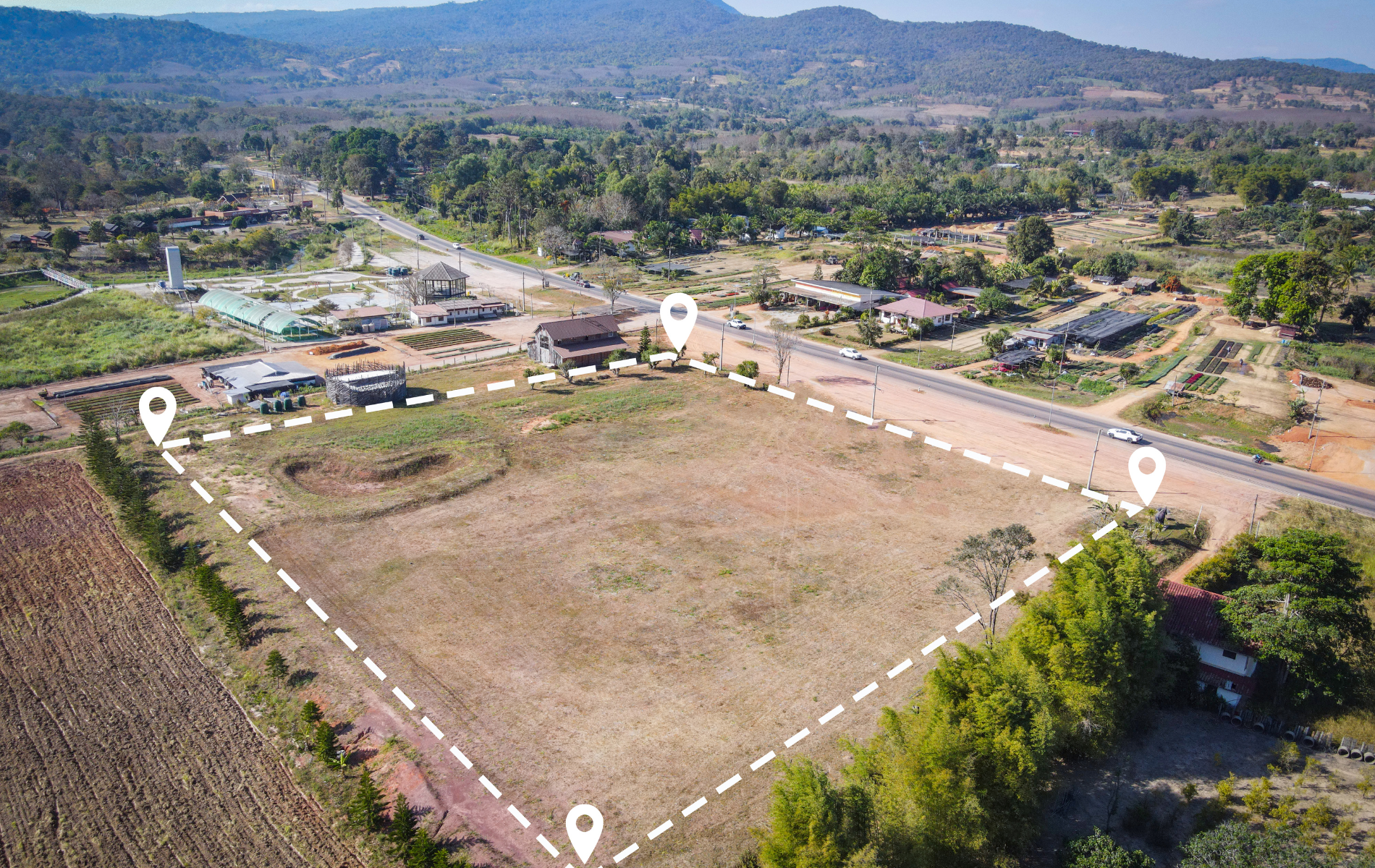 Aerial view of a vacant lot outlined with white dots, near a road and buildings, with trees and a mountain in the background.