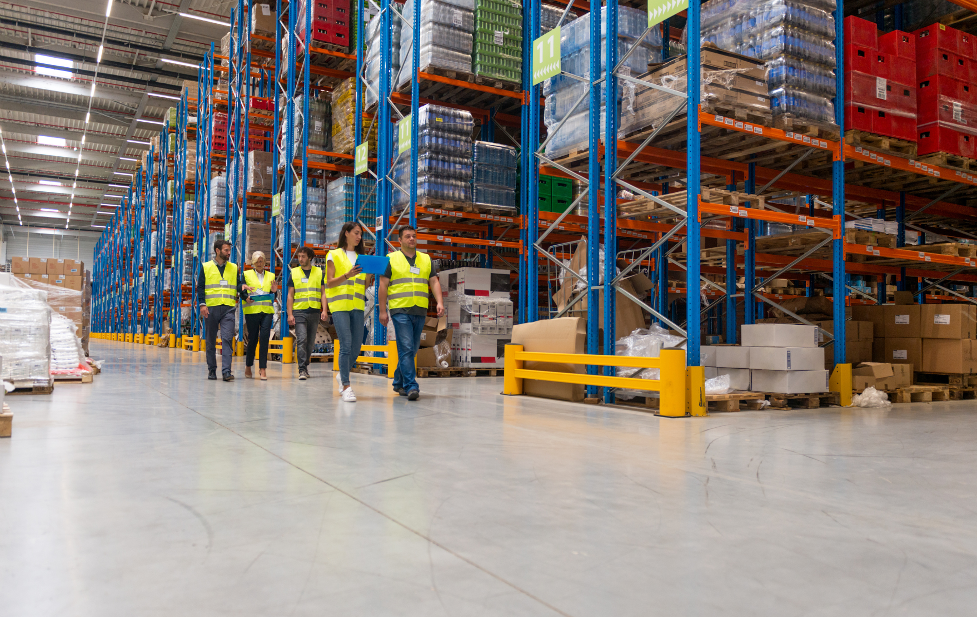 Warehouse workers in yellow vests walk down an aisle with tall shelves of goods.