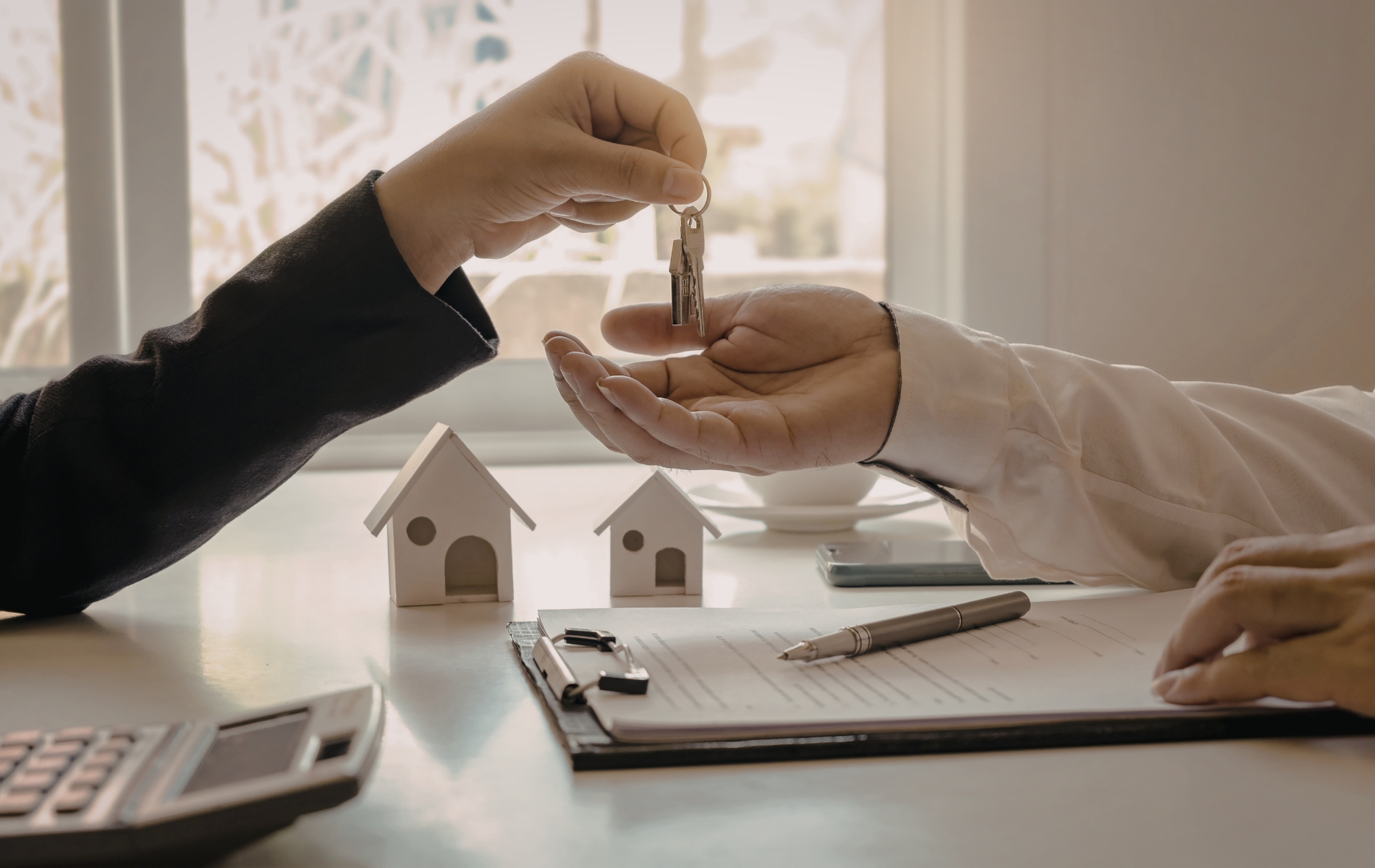 Hands exchanging keys over a table with miniature houses and a document; a real estate transaction.