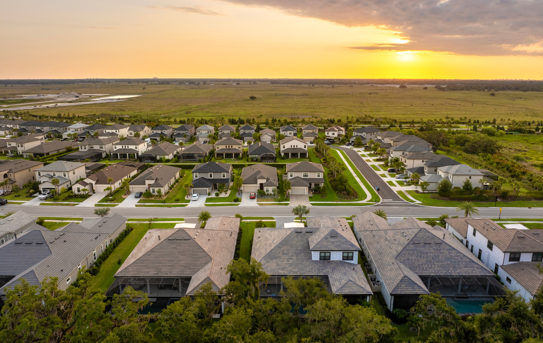 Aerial view of a suburban neighborhood at sunset; houses with green lawns and a field in the distance.