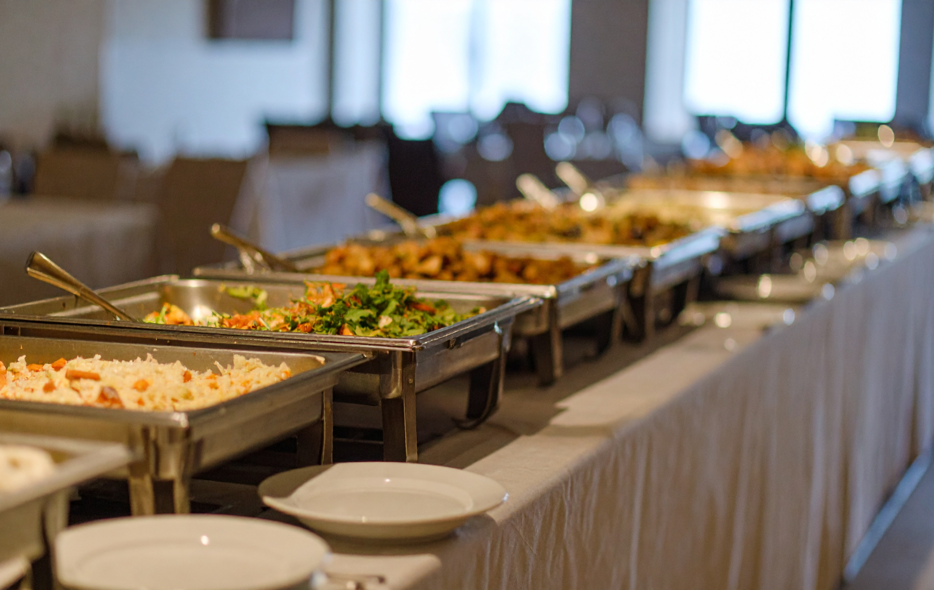 Buffet with various dishes in metal serving trays on a white tablecloth, indoors.