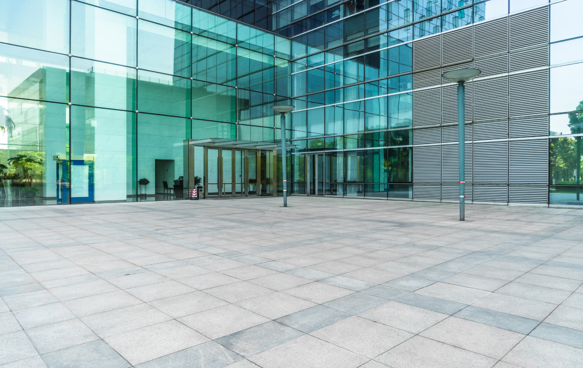 Glass-walled office building entrance with paved plaza; teal and blue glass, gray pavement.