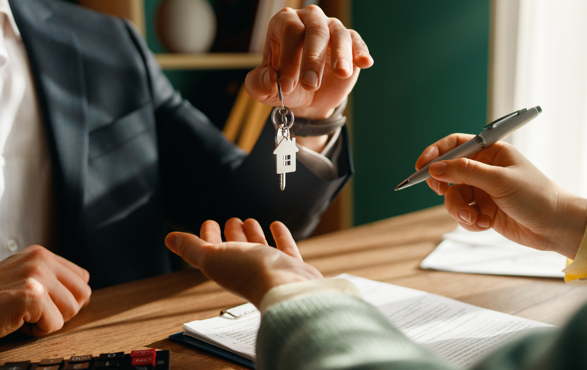 Person handing keys with house-shaped fob to another person over a signed document.