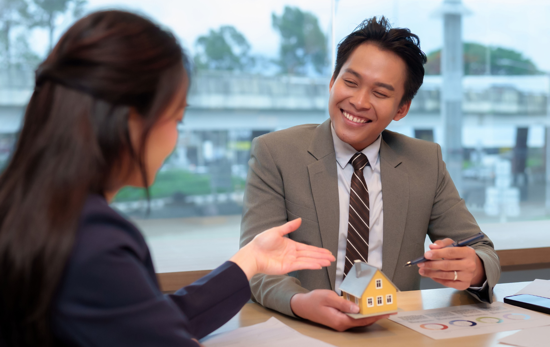 Man in suit smiling, showing a small house model to a woman in an office setting.